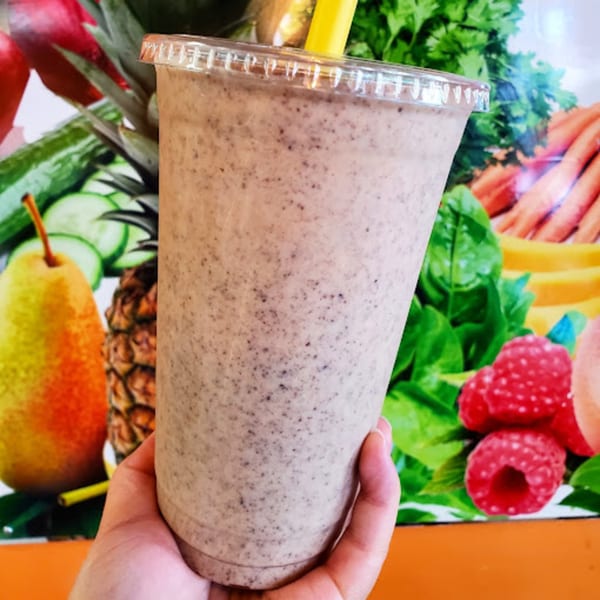 Hand holding a Coco Loco Smoothie with a yellow straw, against a backdrop of colorful fruits and vegetables.